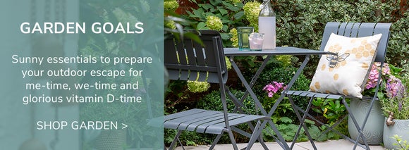 Garden Goals. A 2 seat grey bistro set in a small garden space with drinks on the table and a bee cushion on one of the chairs. Shop Garden >